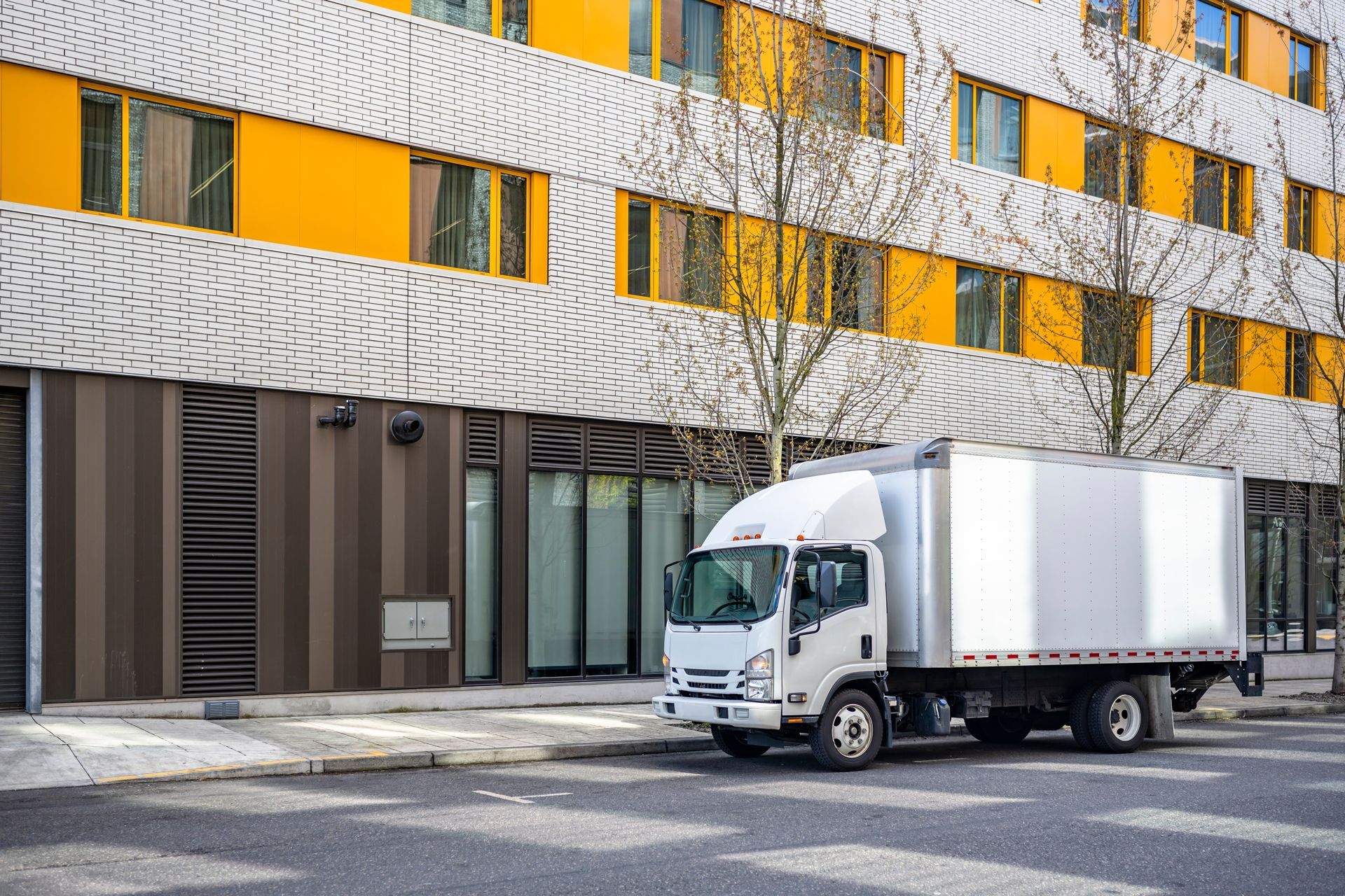 White box truck parked on street in front of modern building with yellow and white facade.