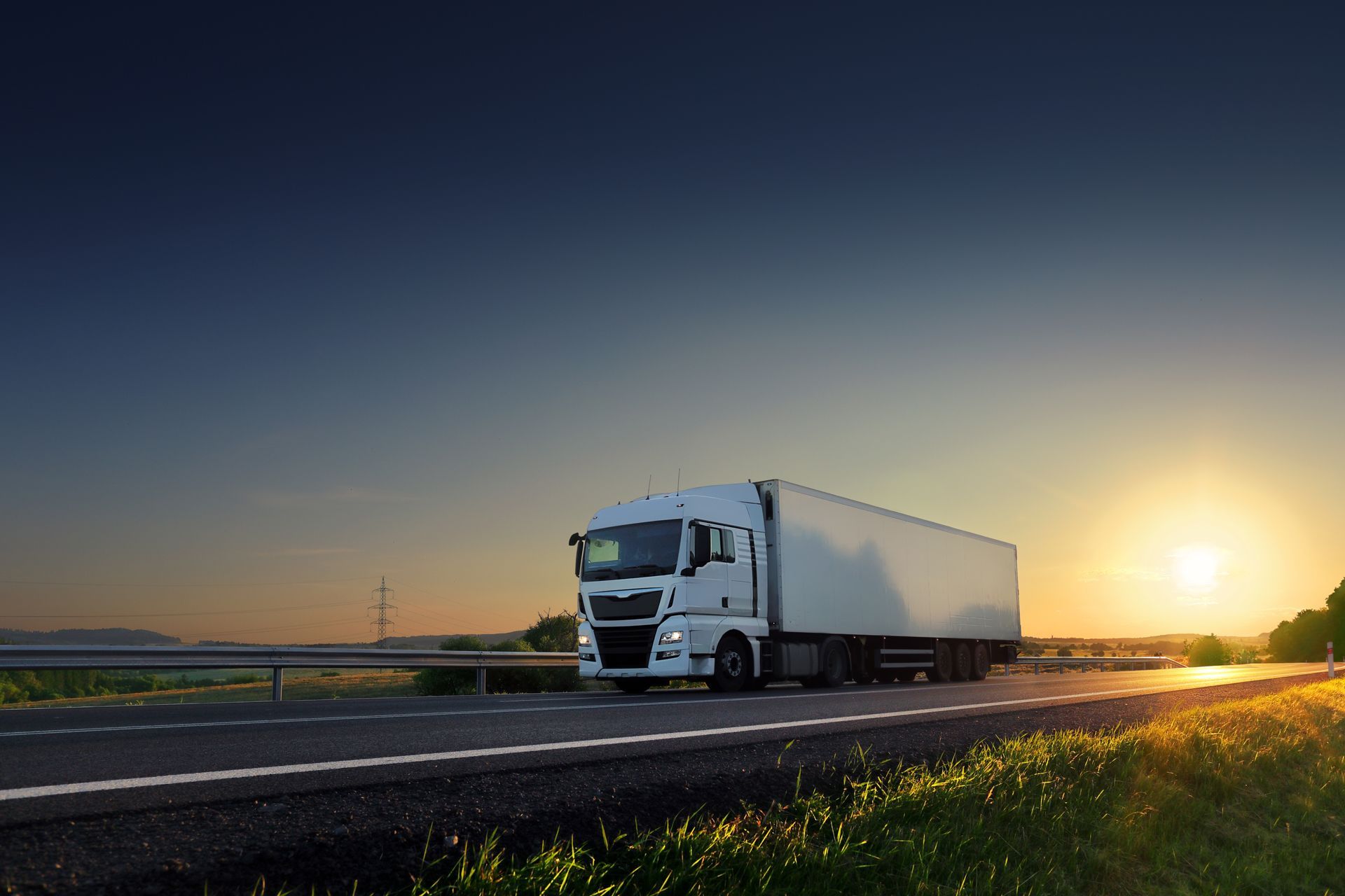 White semi-truck driving on a road at sunset, with a bright sun in the background.