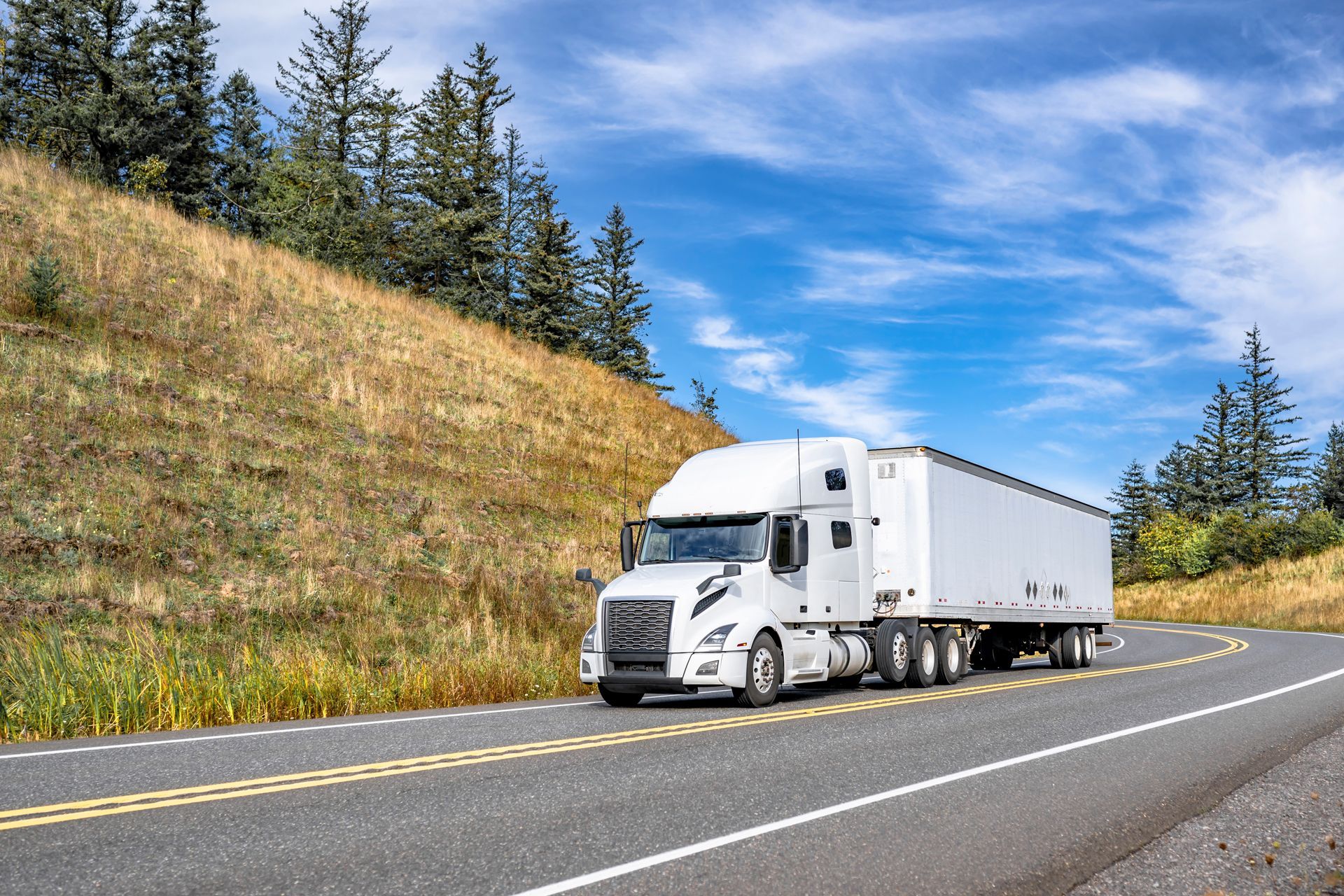 White semi-truck drives on a curved road, with a grassy hill and blue sky in the background.