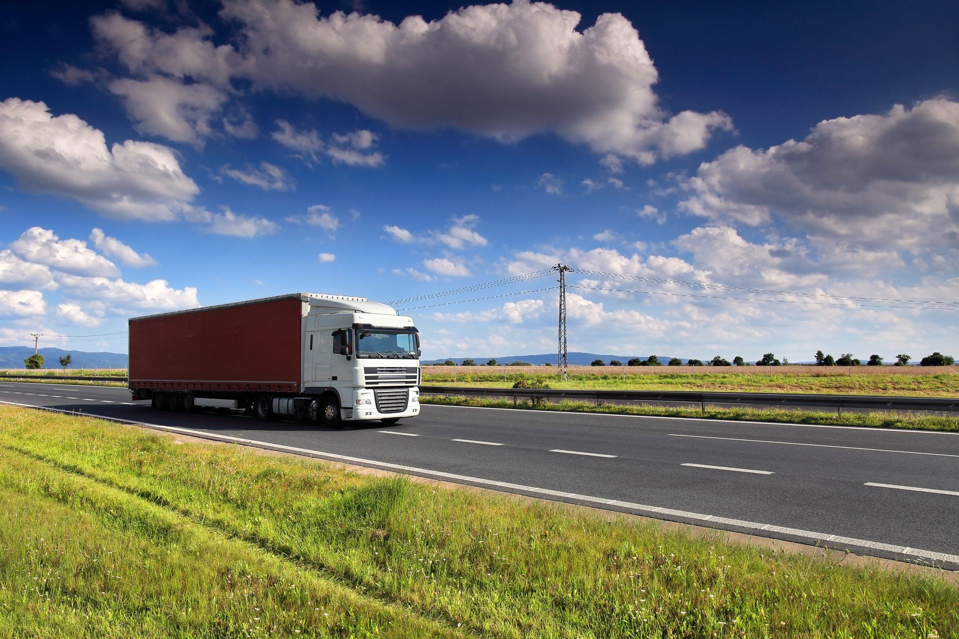 Semi-truck driving on a road, fields of green, blue sky, and white clouds.
