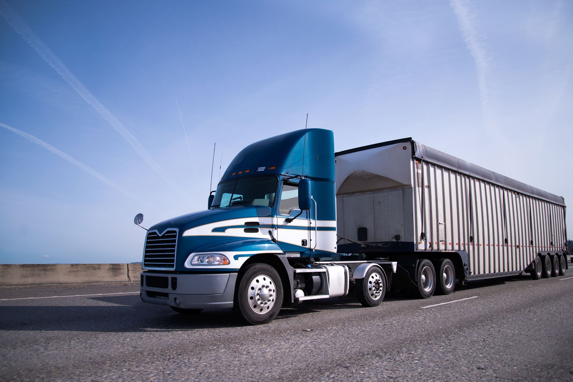 Blue and white semi-truck with trailer on a highway under a blue sky.