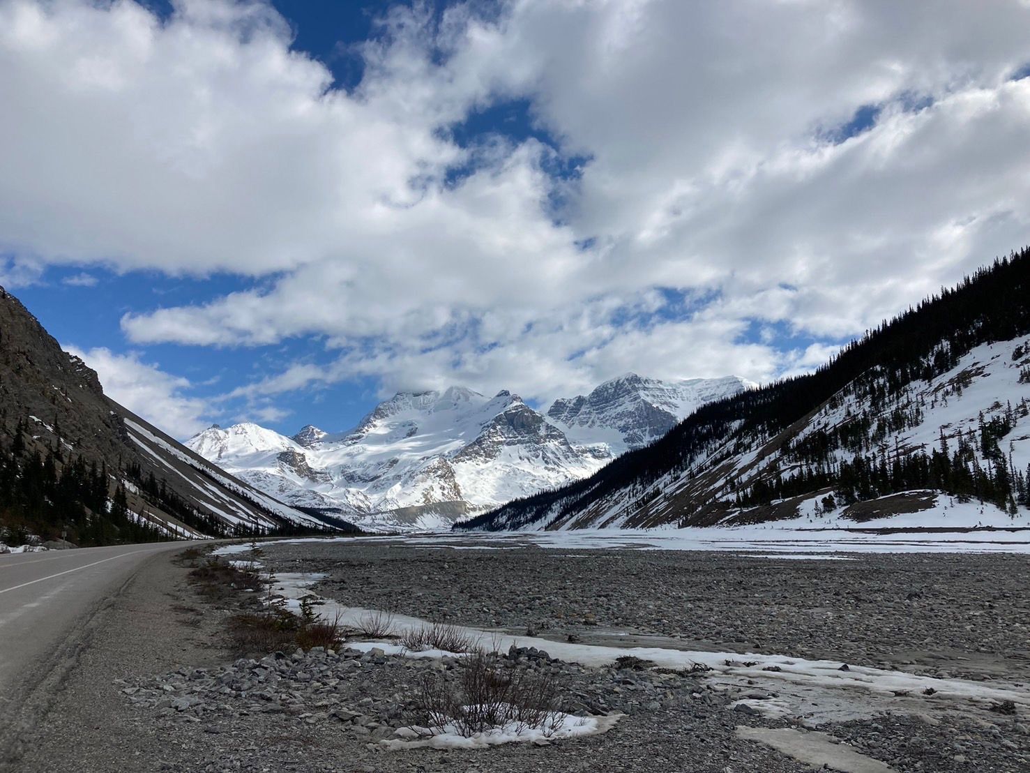 Mountain valley with a river, snow patches, and cloudy sky between dark, snow-dusted slopes