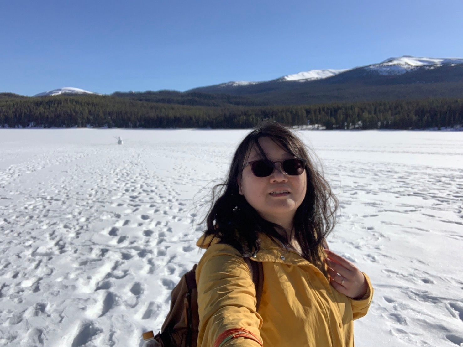 Person in yellow jacket taking a selfie on a snowy lake with mountains in the background
