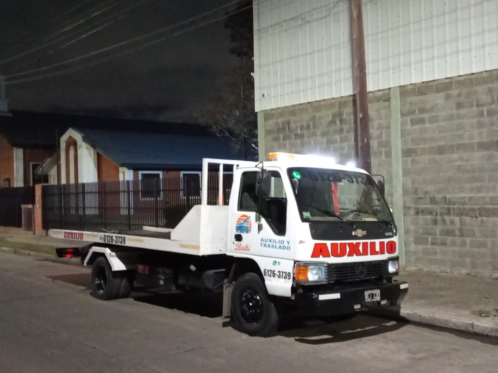 Gr&uacute;a blanca con la palabra &laquo;AUXILIO&raquo; en letras rojas, estacionada en una calle poco iluminada por la noche.