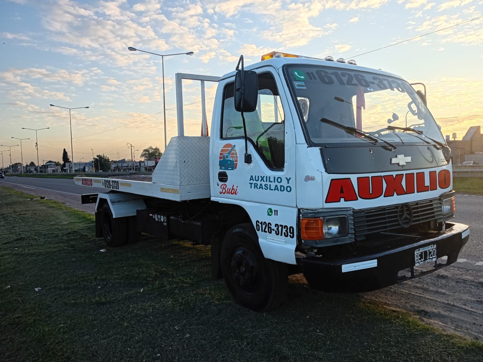 Gr&uacute;a blanca con &laquo;AUXILIO&raquo; aparcada a un lado de la carretera al atardecer.