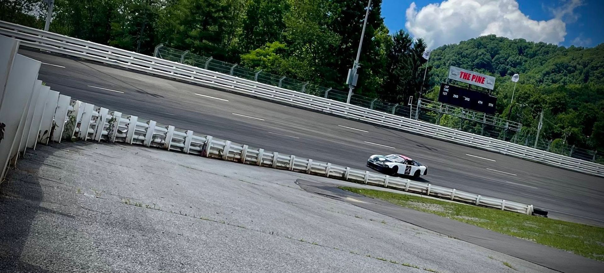 Tilted view of a race track with a white car on the turn and a green hillside in the background