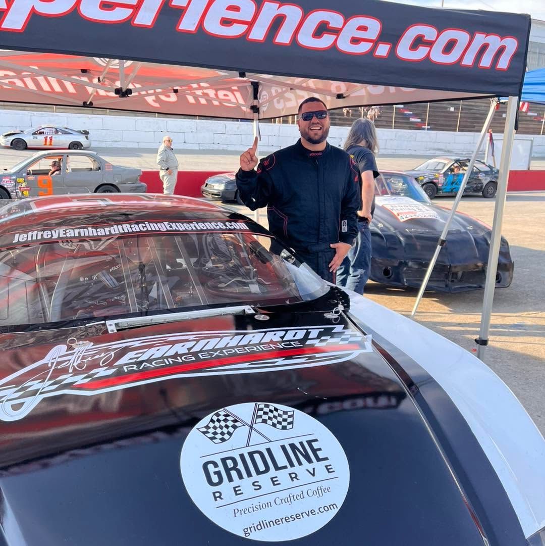 Man posing beside a red race car under a canopy at an авто event, with a Gridline logo on the hood