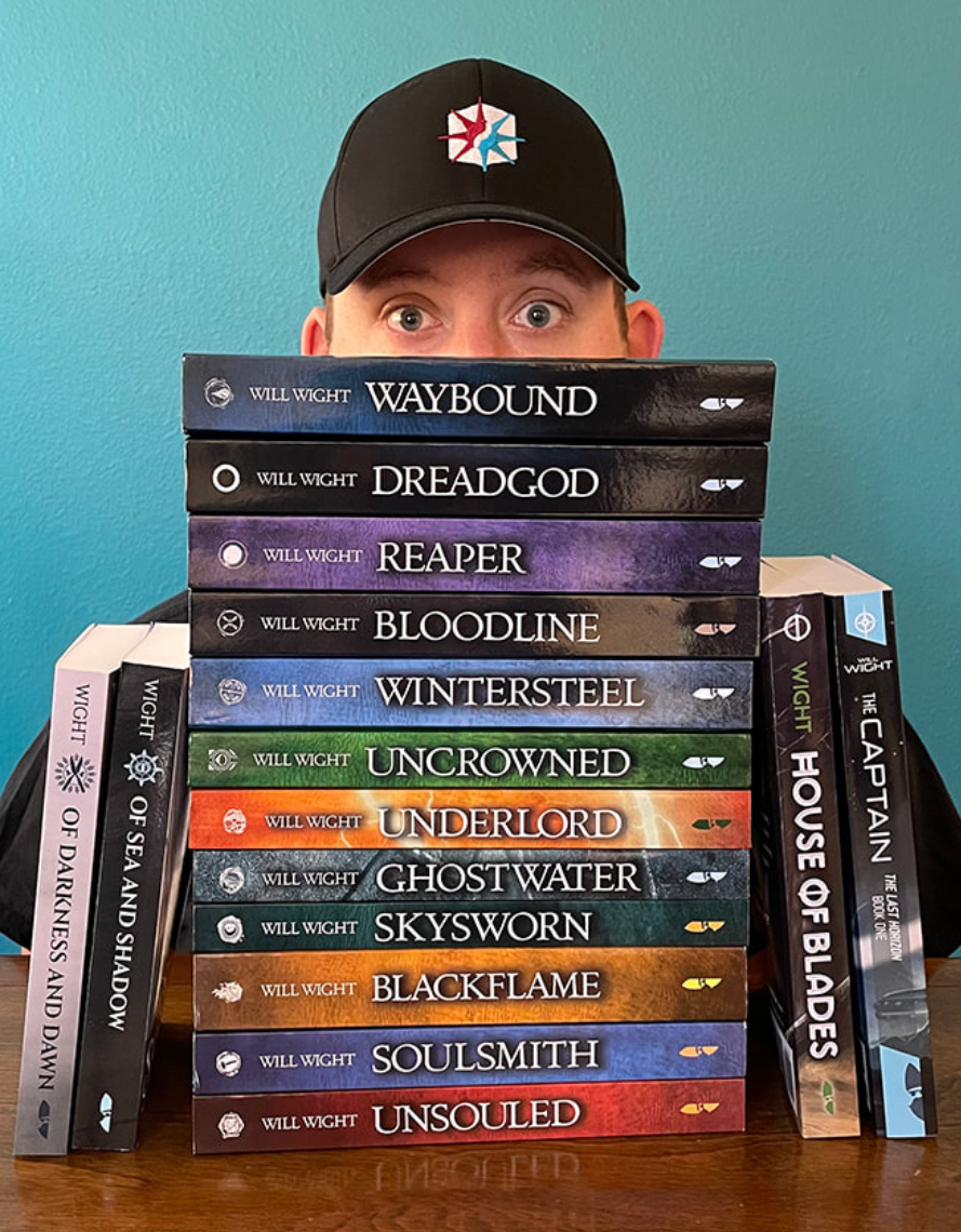Person in black cap peeks over stacked fantasy novels on a wooden table against a teal wall.