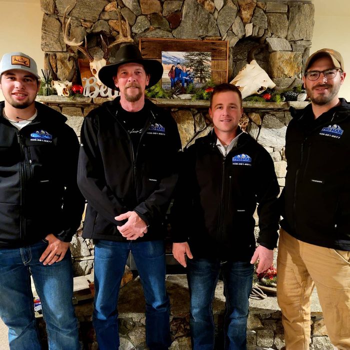 Four men in black jackets and jeans stand in front of a stone fireplace.