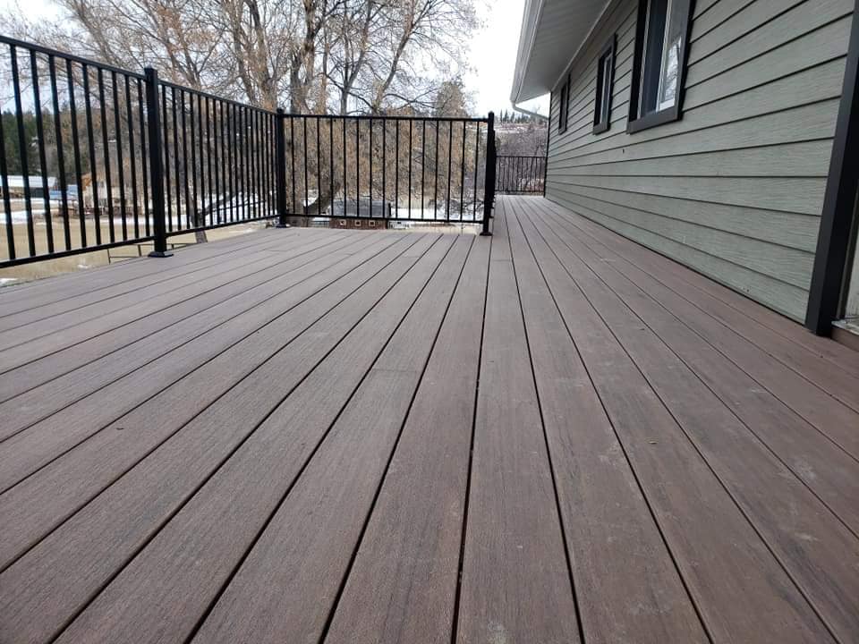 Wooden deck with black railing next to a house with green siding, overlooking a snowy landscape.