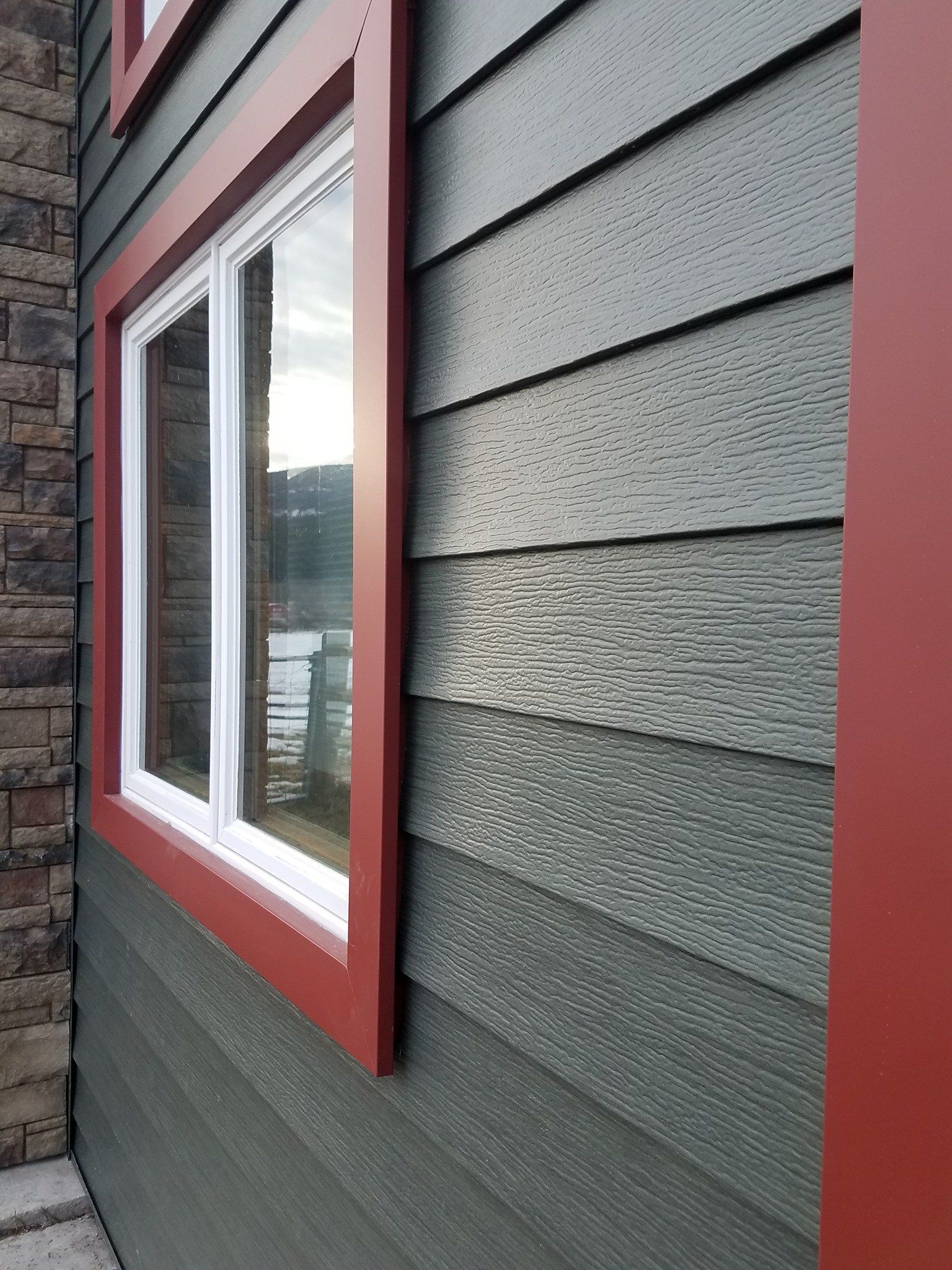Window with red trim on a house with dark green siding next to a stone wall.
