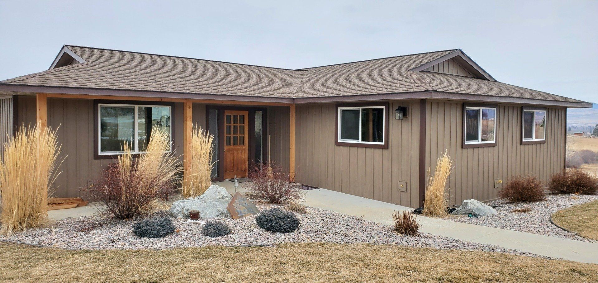 Brown house with brown roof, front landscaping with rocks and plants, cloudy sky.