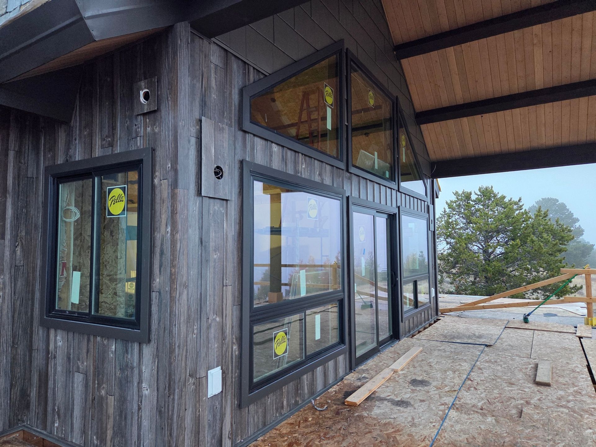 Exterior of a cabin with dark framed windows and weathered wooden siding, partially built with a deck.