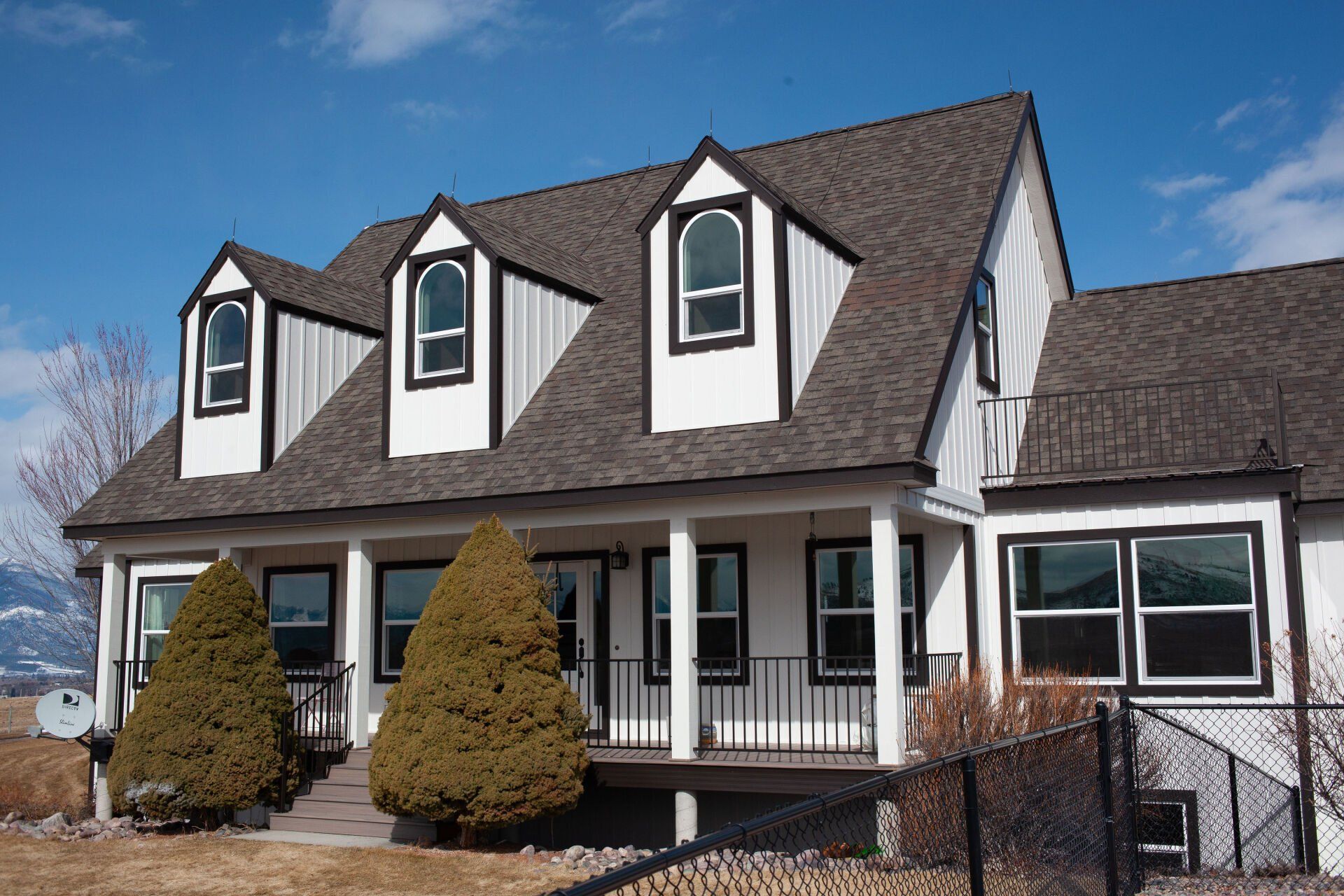 White house with black trim, three dormers, and a porch, set against a blue sky.