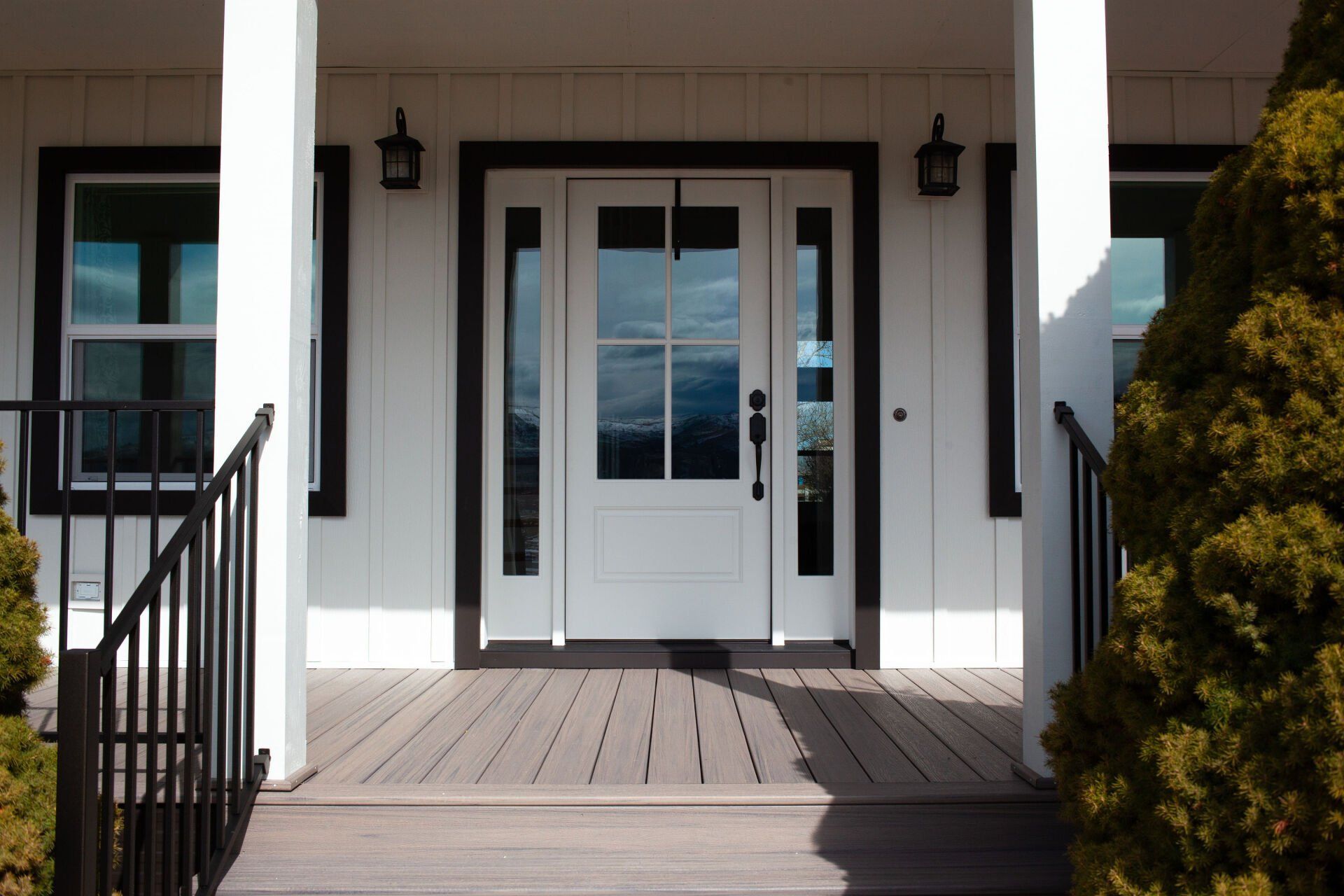 White farmhouse entrance with black trim, windows, and door. Gray porch with a black railing and bush on right.