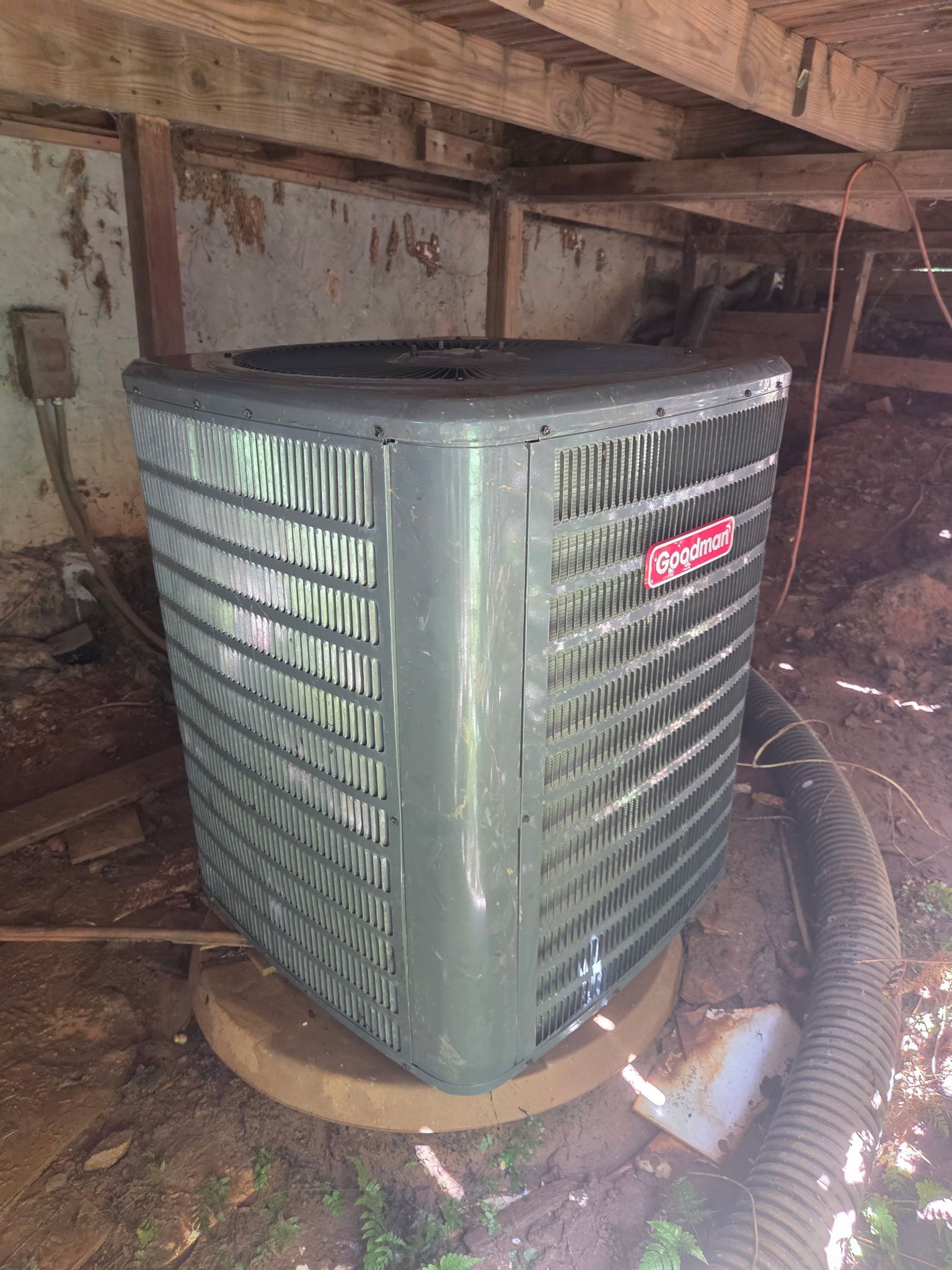 Green outdoor air conditioner unit under a wooden structure, with dusty hose and debris around it