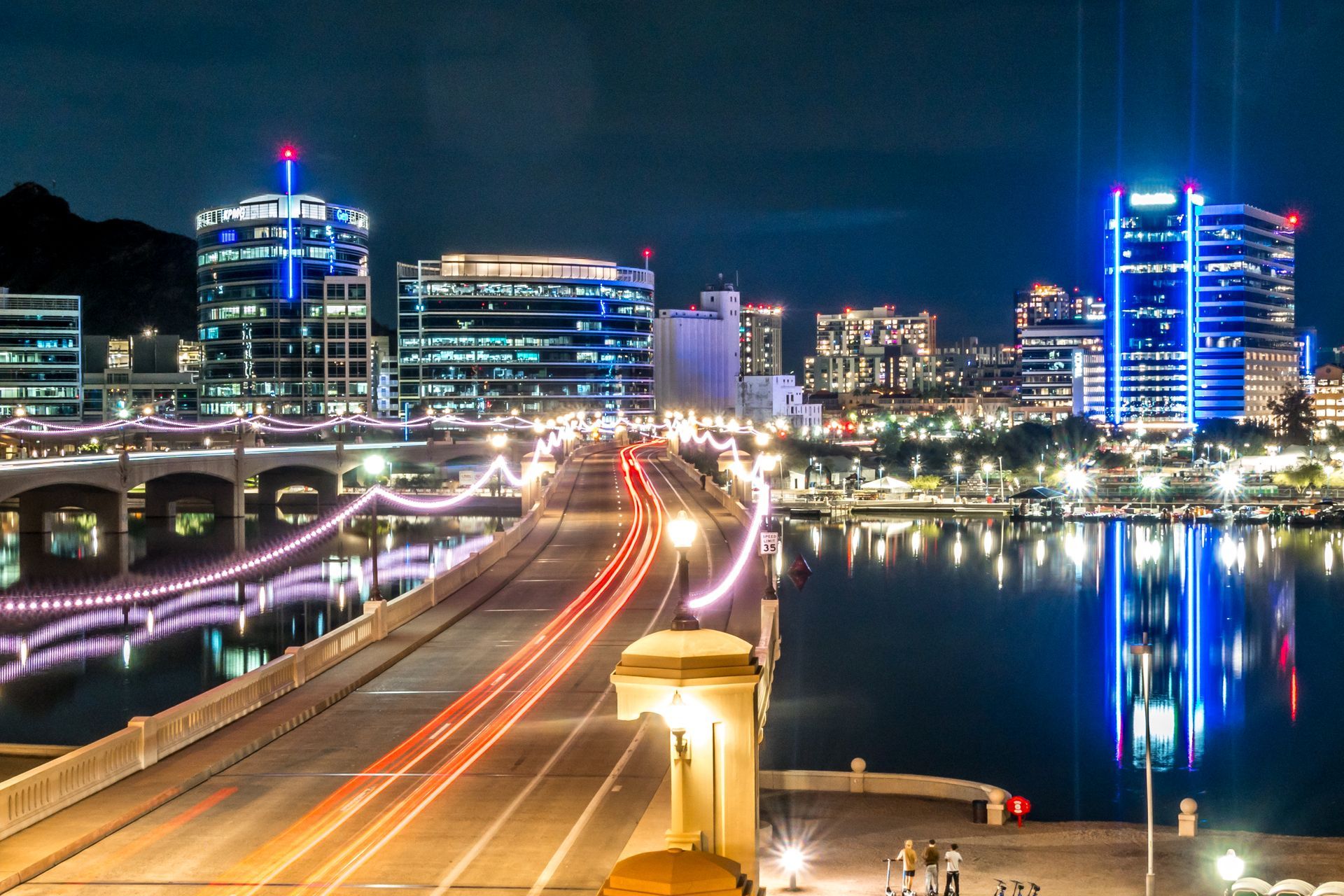 Nighttime city skyline with illuminated buildings, bridge light trails, and reflections on the water.