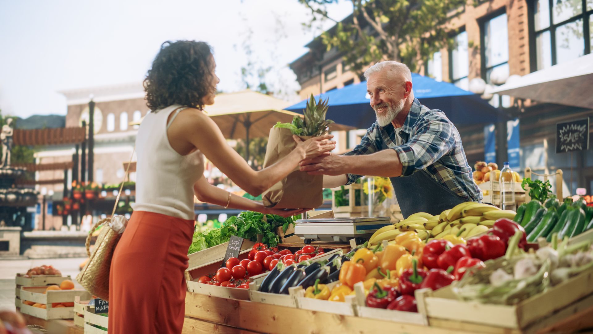 Two people exchanging fresh produce at an outdoor farmers market, with colorful fruits and vegetables displayed.