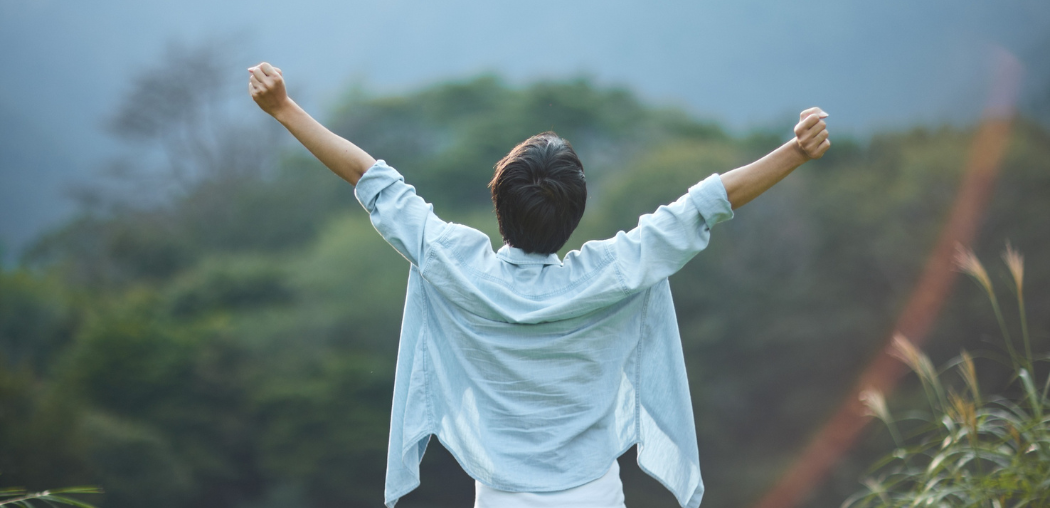 Person in a white shirt stands with arms raised, facing misty green hills.