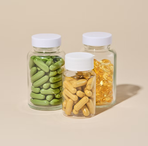 Three small glass supplement bottles with white caps, filled with green, yellow, and amber capsules on a beige background