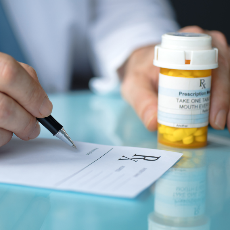 Hand writing a prescription beside a yellow pill bottle on a glass table