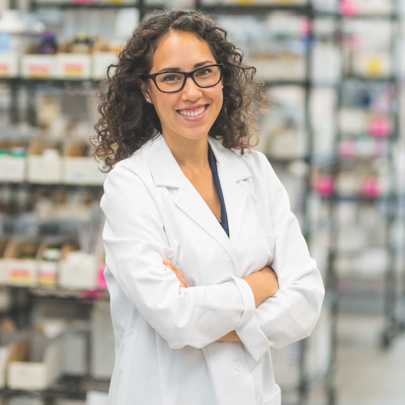 Smiling pharmacist in a white coat with folded arms, standing in a pharmacy aisle.