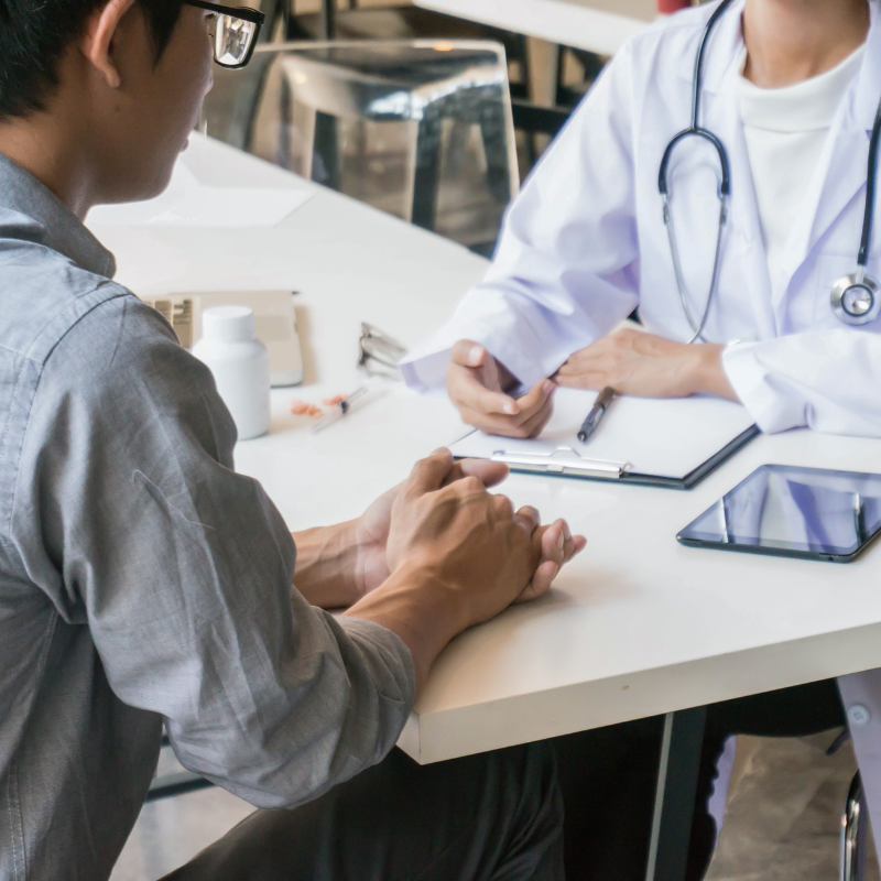 Doctor speaking with patient at a clinic desk, with a tablet and clipboard visible.