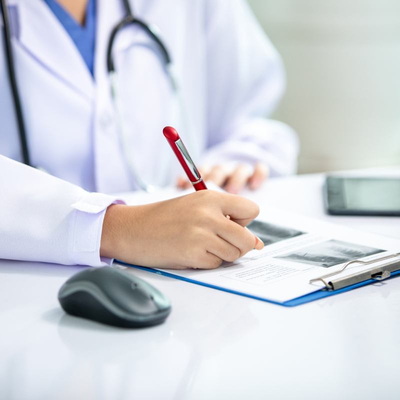 Doctor in white coat writing on a clipboard beside a computer mouse at a desk
