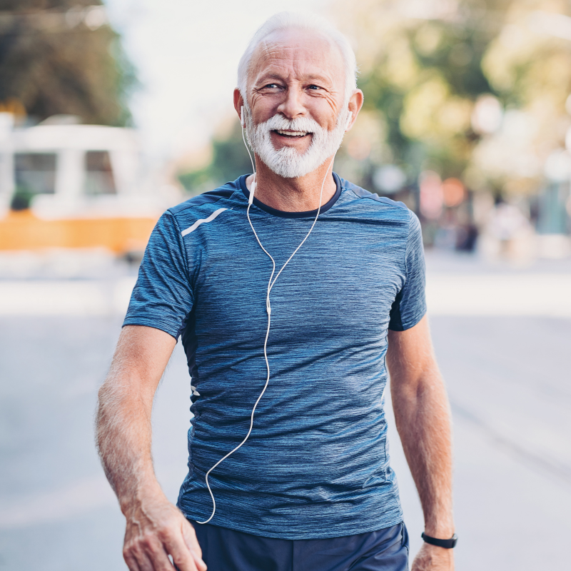 Smiling man in blue running shirt and shorts jogging outdoors with earbuds in a sunlit park