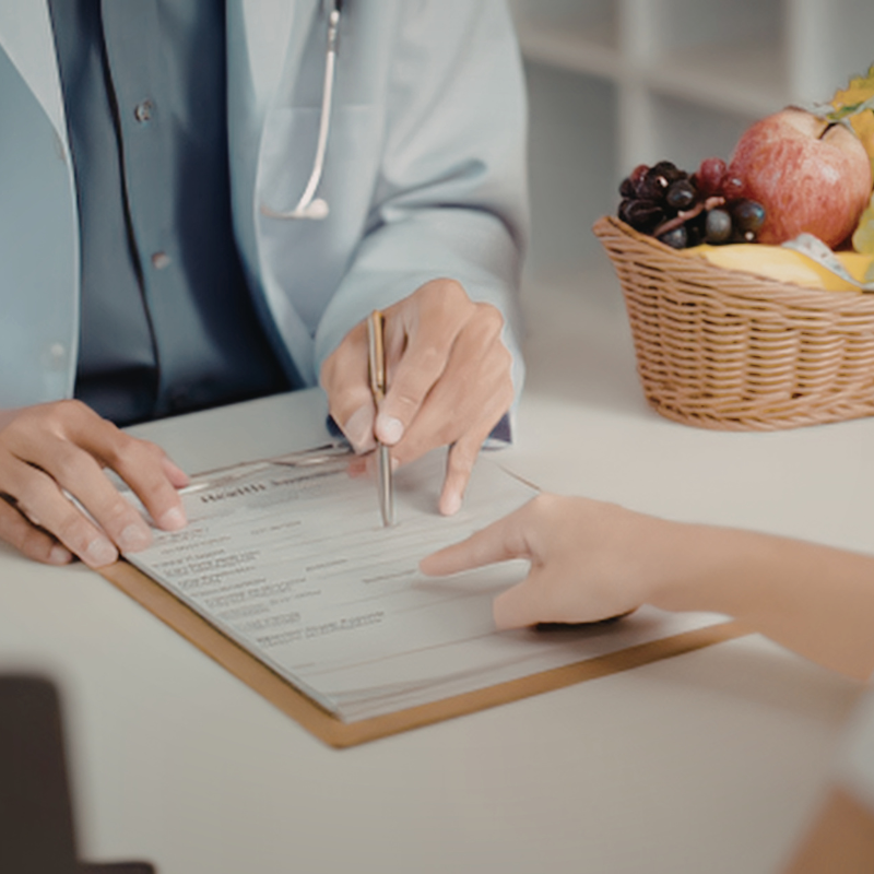 Person signing a document at a desk beside a fruit basket, with another hand pointing to the form.