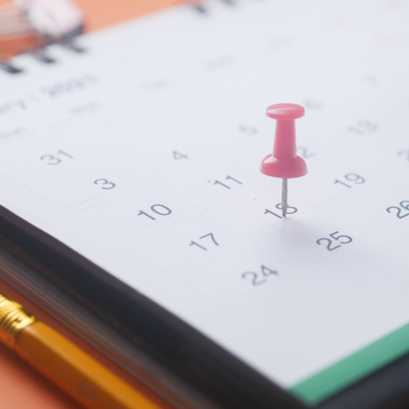 Pink pushpin marking a date on a paper calendar beside a gold pen