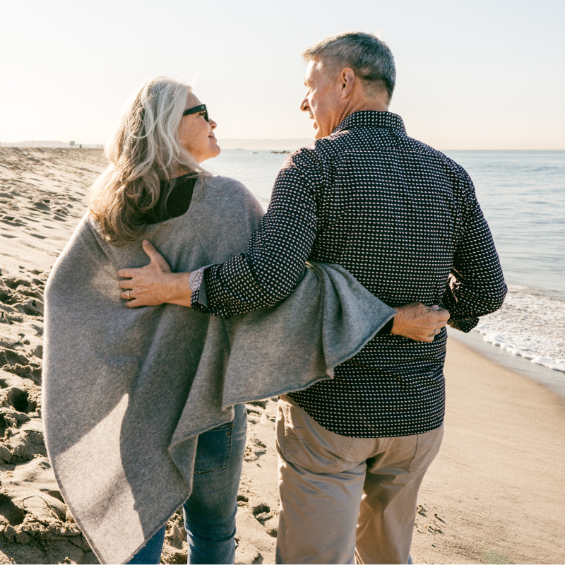 Two people walking arm in arm on a sunny beach, facing the ocean