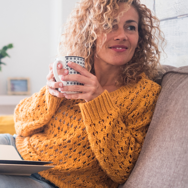 Woman in a mustard sweater holding a mug on a couch, smiling by a bright window