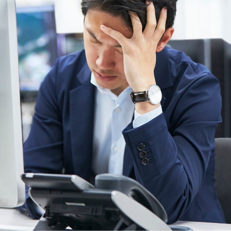 Stressed office worker at desk, holding his forehead beside a desk phone and computer.