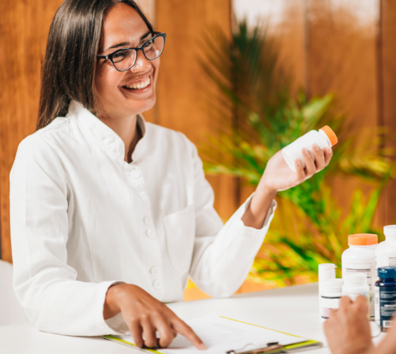 Smiling person in a white coat holding a pill bottle at a lab table with medical containers