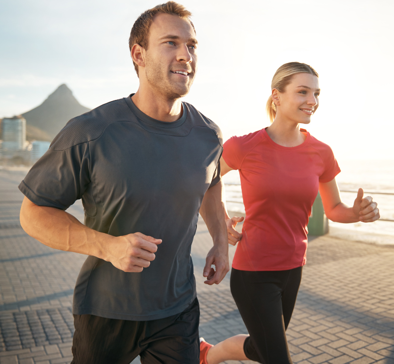 Two runners jogging on a sunlit waterfront path, with a city skyline in the background