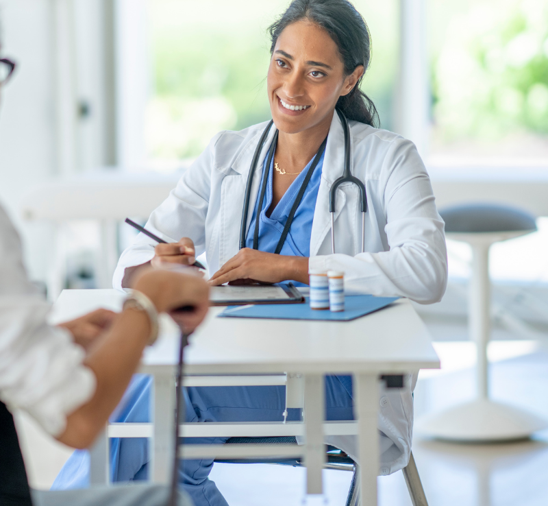Doctor smiling at a desk with a stethoscope, writing notes during a consultation.