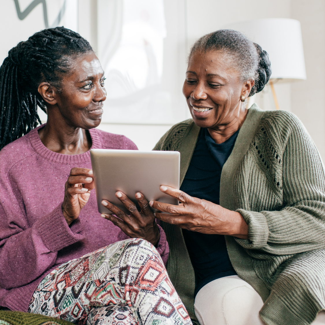 Two people smiling and sharing a tablet on a couch in a bright living room