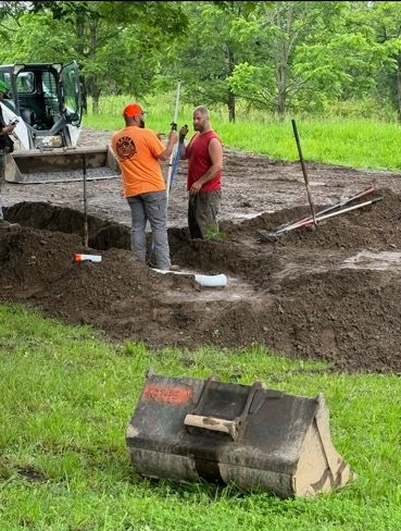 An excavator bucket lifts dirt next to a large, beige underground storage tank in a residential yard.