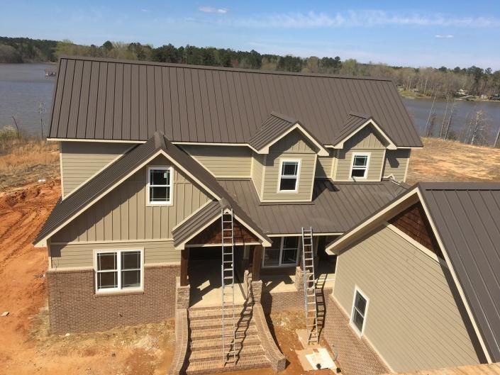 Aerial view of a new tan house with a dark roof, front entry, and surrounding dirt lot near water.