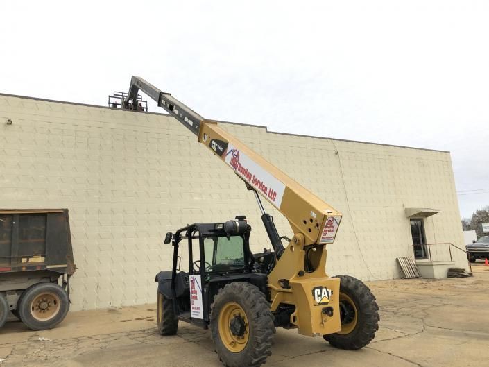 Yellow CAT telehandler extending boom to a building roof beside a parked trailer in a dusty lot