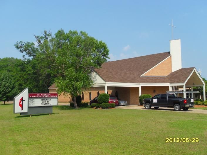 Small brick church with a white steeple, sign, trees, and parked vehicles on a grassy lot