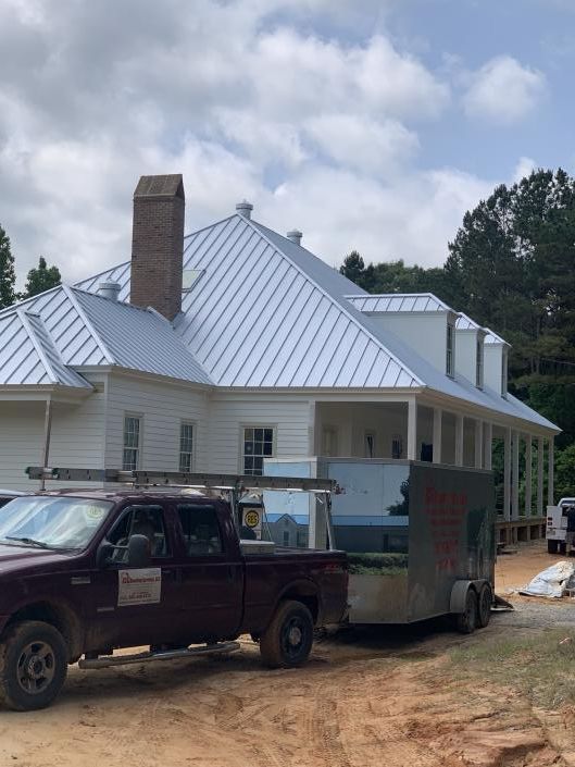 White house with metal roof and brick chimney, with a black pickup truck and silver trailer in front