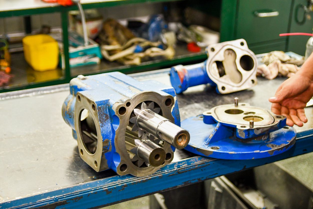 Metal gearbox parts on a workbench in a workshop, with a person’s hand reaching in from the right.