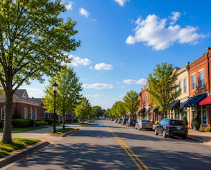 Sunny small-town street with brick shops, parked cars, and leafy trees under a blue sky