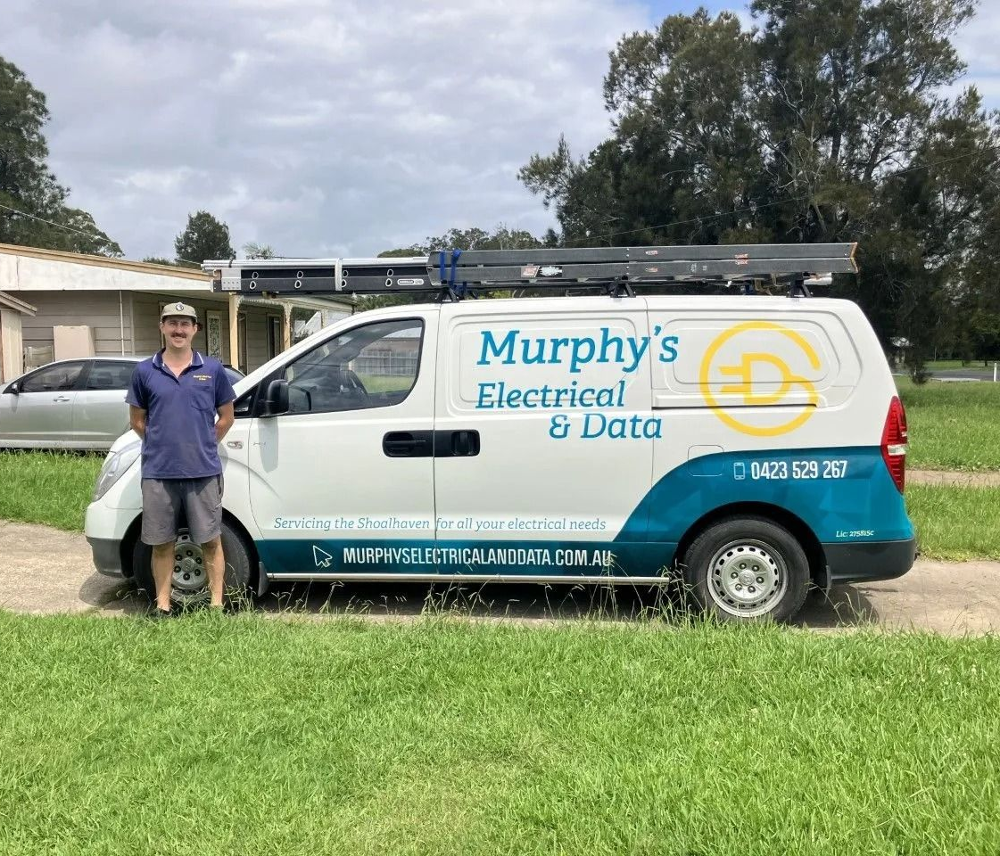 Man standing by Murphy's Electrical & Data van; white van with teal accents; green grass, trees in the background.