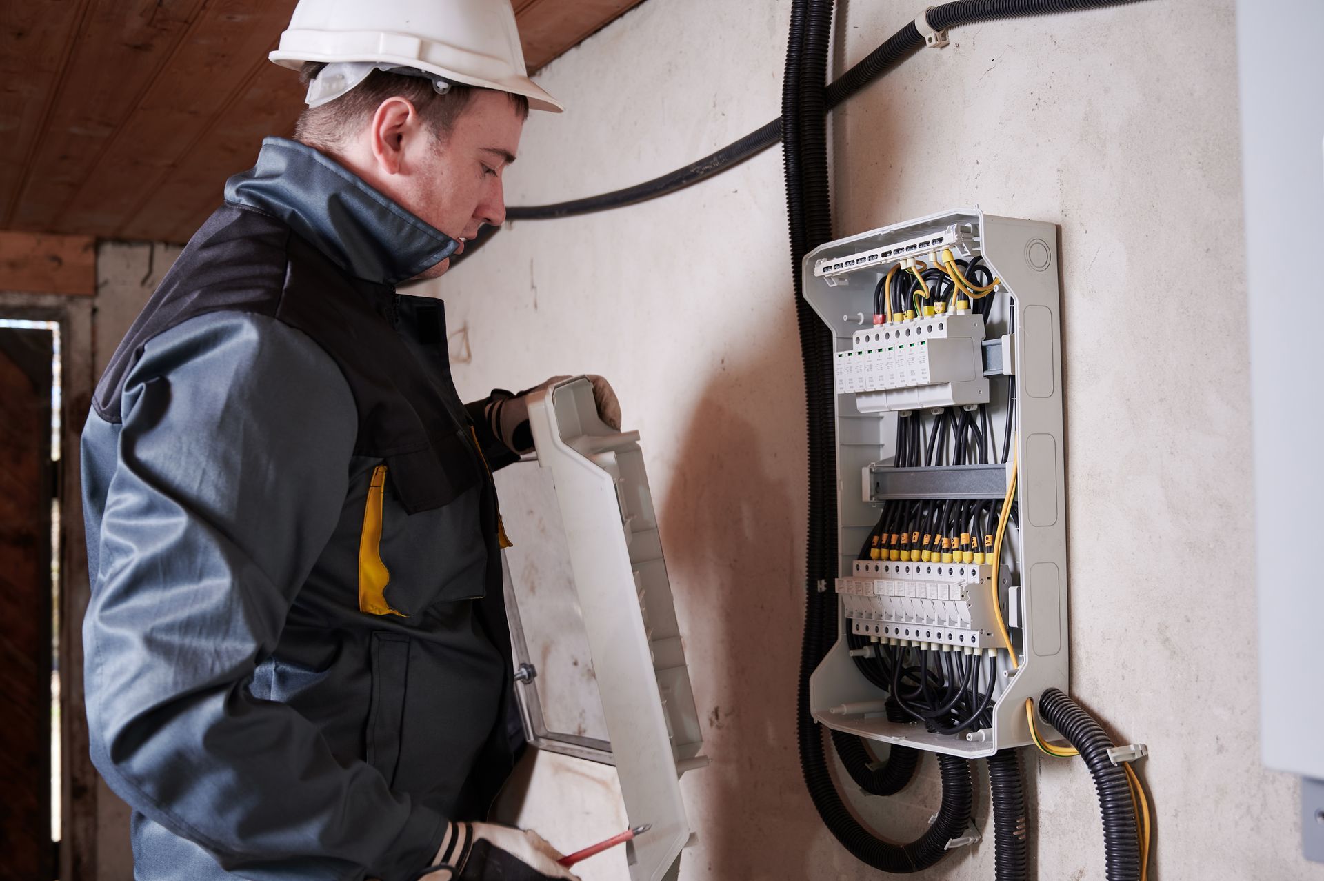 An electrician wearing a white hard hat and work jacket inspects an open electrical panel mounted on a wall.