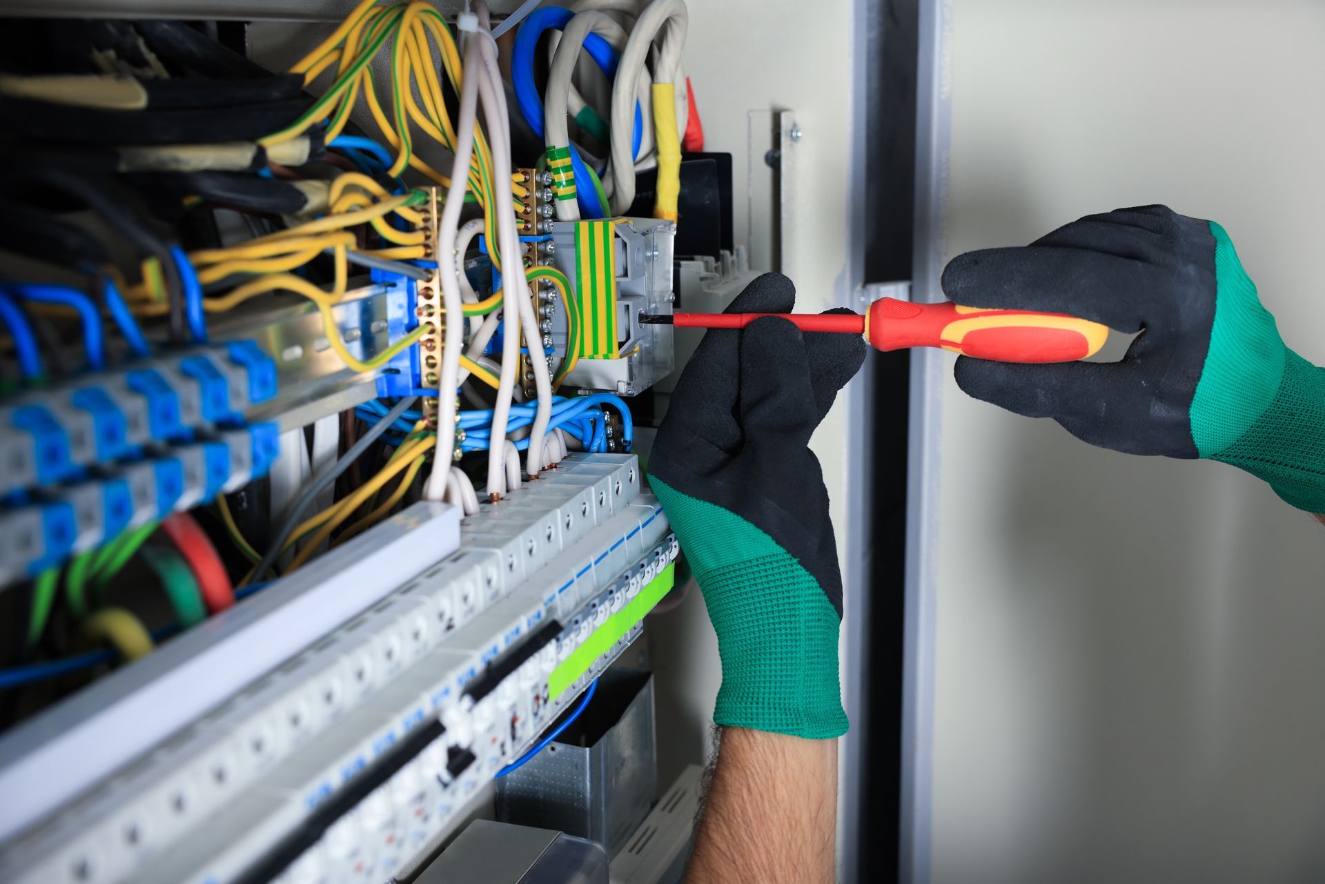 An electrician in safety gloves uses a screwdriver to tighten a connection in a complex electrical control panel.