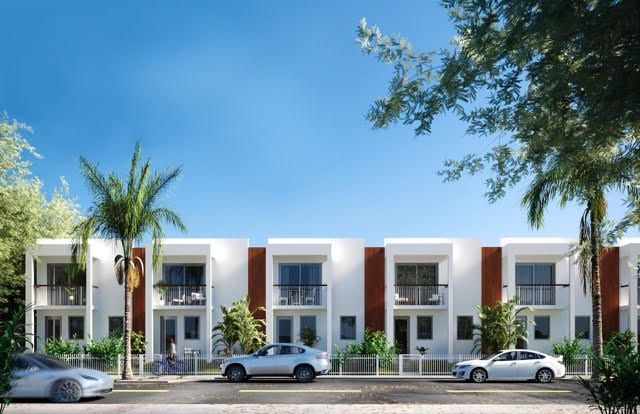 Modern white townhouses with balconies and parked cars along a sunny street, framed by palm trees.