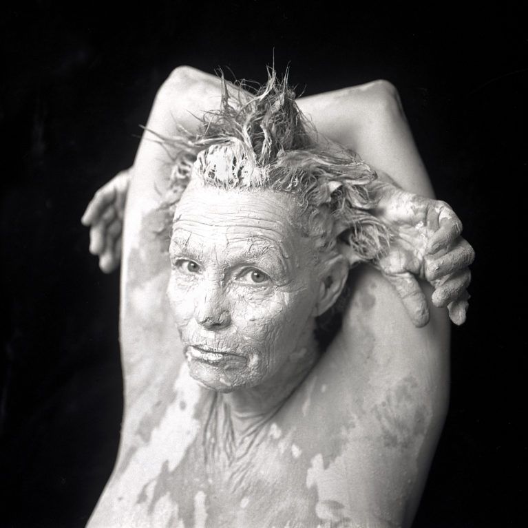 Person covered in white cream with arms raised and hair spiked, looking at the camera against a black background